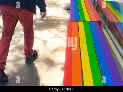 Homme marchant sur un trottoir avec banc en bois dans les couleurs de l'arc en ciel, symbole de la fierté gay Banque D'Images