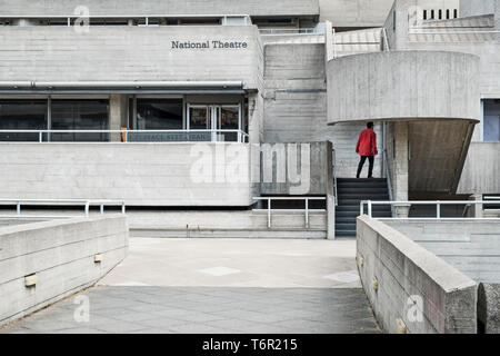 Londres, Royaume-Uni. Le Théâtre National, par l'architecte Denys Lasdun (1977). Un homme portant une veste rouge vif se détache sur les surfaces de béton gris Banque D'Images