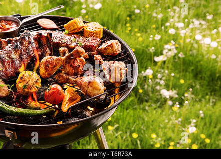 Cuisson barbecue d'été plus d'un feu chaud avec légumes variés, T-bone steak et cuisses de poulet épicé sur le grill grésillant dans un pré vert Banque D'Images