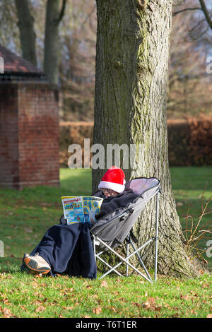 Enfant isolé dans le parc par arbre portant un chapeau de Santa, assis dans une chaise de camping, enveloppé dans une couverture en polaire, souriant et lecture de bande dessinée en hiver soleil. Banque D'Images