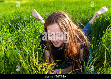 Jeune femme de race blanche aux cheveux rouges avec des taches de rousseur, reposant sur un champ vert au coucher du soleil de printemps. Banque D'Images