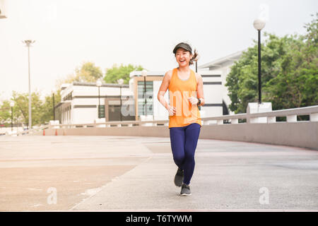 Une jeune femme asiatique et active qui porte un chapeau pare-soleil arrière et t-shirt jaune, elle s'exécute dans un parc public avec un visage heureux et sourire Banque D'Images