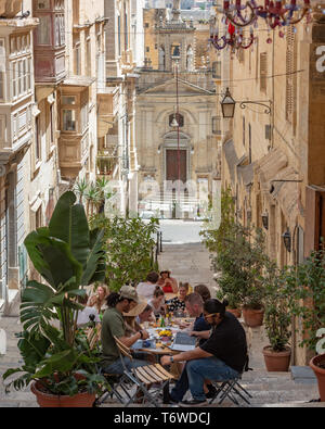 Les amis apprécient un déjeuner en plein air dans un restaurant situé dans l'étroite rue Sainte-Lucie de la Valette, avec l'église Sainte-Lucie en arrière-plan. Banque D'Images