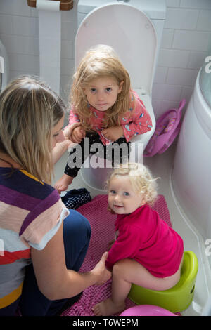 Les jeunes enfants dans les toilettes avec leur mère, on s'assoit sur les toilettes, alors que sa jeune sœur utilise un petit pot. Banque D'Images