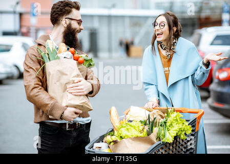 Young couple with shopping bags et panier plein de produits frais sur le parking près du supermarché outdooor Banque D'Images