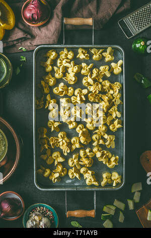 Des pâtes tortellini végétarien cru dans le plateau métallique sur fond foncé table de cuisine rustique avec des ingrédients et des ustensiles, vue d'en haut. La cuisson prepar Banque D'Images