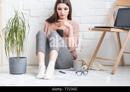 Une femme heureuse dans des vêtements décontractés est assis dans un intérieur lumineux et utilise un téléphone, la jeune fille se repose en travaillant sur un ordinateur portable et le surf Banque D'Images