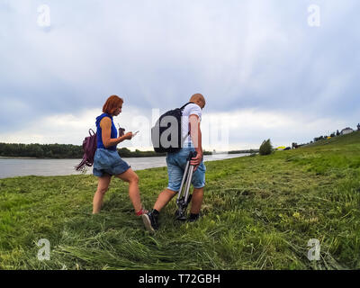 Le gars et la fille en short et T-shirts aller en haut de l'herbe bien verte sur la rive du fleuve avec des sacs à dos. Une femme a un café et un téléphone dans les mains, Banque D'Images