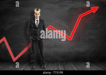 Vue avant pleine longueur de businessman wearing crown, debout les mains dans les poches, avec flèche graphique rouge qui monte derrière lui à black graph-gouverné wall. Banque D'Images