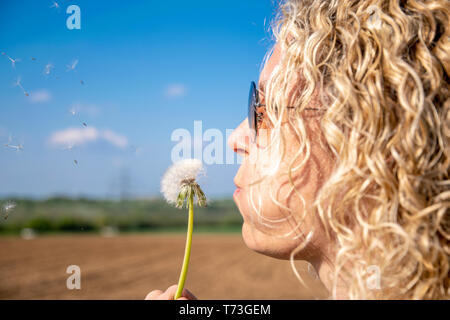 Une blonde woman blowing dandelion en plein essor et les graines s'envolent de ciel bleu Banque D'Images