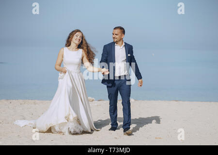 Belle jeune femme aux cheveux longs en robe blanche avec son jeune mari sur la plage Banque D'Images