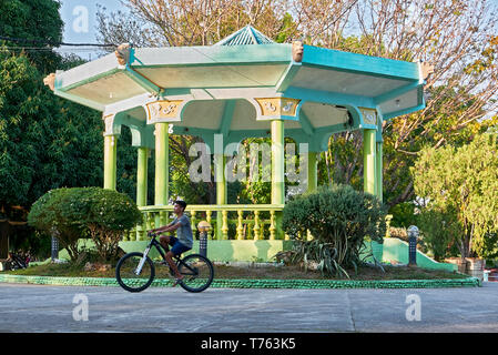 Leon, Iloilo, Philippines : Teenage boy riding a bike en face d'un pavillon de couleur verte à la ville plaza park dans l'heure d'été Banque D'Images