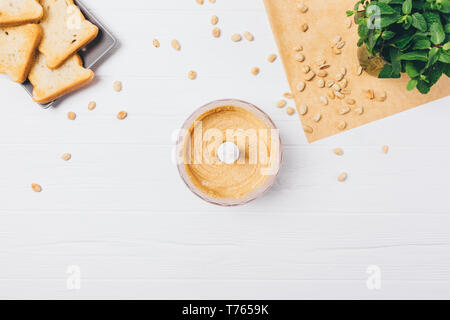 La cuisson de beurre d'arachide croquant frais ; propagation dans blender bol à côté de pain grillé sur une table de cuisine en bois blanc, vue du dessus. Banque D'Images