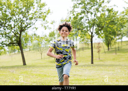 Happy fit jeune garçon courir vers l'appareil photo dans l'herbe verte dans un jardin de printemps avec un grand sourire amical Banque D'Images