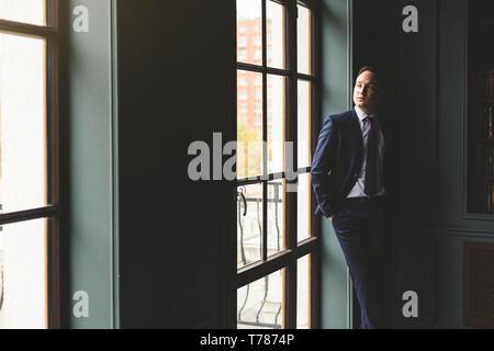 Portrait d'un homme debout dans un costume d'affaires coûteux près de la fenêtre de son bureau et sourit. Banque D'Images