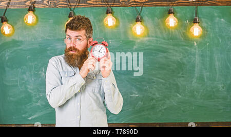 Enseignant dans les lunettes est titulaire d'un réveil. Homme avec barbe et moustache sur le visage concentré d'horloge d'écoute. Cloche de concept. Hipster barbu est titulaire d'réveil, tableau sur toile de fond, l'espace de copie. Banque D'Images