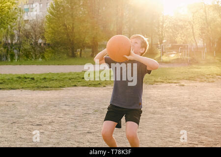Jeune garçon jouant au basket-ball avec pour objectif de prendre la balle à l'objectif avec un regard de la concentration d'un rétro-éclairage par flare du soleil du soir Banque D'Images