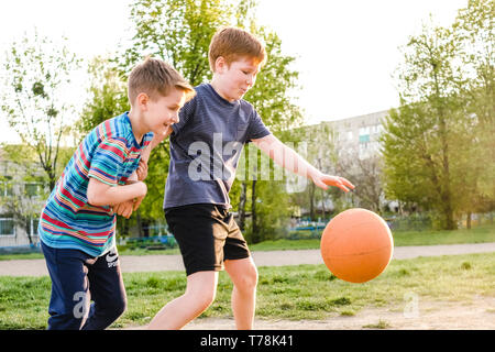 Deux jeunes garçons à la poursuite d'un basket-ball tout en pratiquant leur jeu à l'extérieur, sur un terrain de jeu dans la douce lueur du soleil Banque D'Images