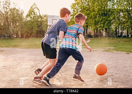 Deux jeunes garçons à la poursuite d'un basket-ball tout en pratiquant leur jeu à l'extérieur, sur un terrain de jeu dans la douce lueur du soleil Banque D'Images