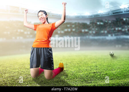 Joueur de football asiatique femme célébrer son but avec les bras levés et à genoux sur le terrain de football au stade Banque D'Images