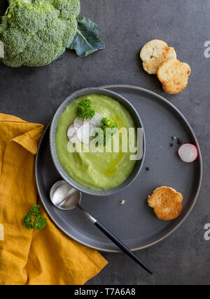 Crème de brocoli soupe avec du pain et une cuillère de radis sur plaques gris, avec serviette orange sur fond gris. Le brocoli cru frais près de. Concept de foo en bonne santé Banque D'Images