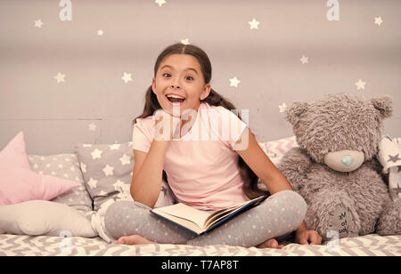 Deviner de son conte de fées. Petite fille s'asseoir lit avec ours lire livre. Kid se préparer à aller au lit. Temps agréable dans une chambre confortable. Kid fille cheveux longs pyjamas mignons vous détendre et lire livre pour porter jouet. Banque D'Images