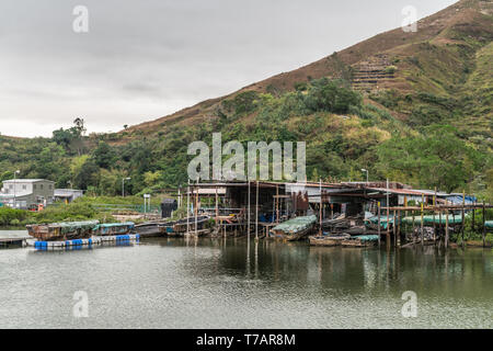 Hong Kong, Chine - mars 7, 2019 : les pêcheurs des maisons sur pilotis sur Tai O Rivière avec grande colline verte en arrière sous ciel gris. Sloops, bâches et dwe simple Banque D'Images