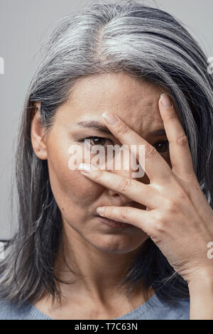 Belle femme mature Asian Gray-Haired avec la peau foncée et les yeux bruns couvre le visage avec sa main et regarde vers la caméra. Sa tête inclinée. M naturel Banque D'Images