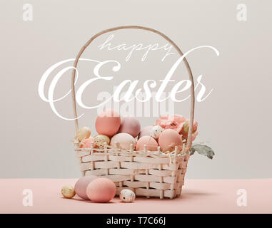 Panier en osier avec des oeufs peints rose, fleurs et joyeuses Pâques des lettres blanches sur fond gris Banque D'Images
