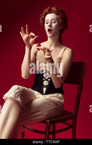 Jeune femme rousse médiévale comme une duchesse en noir corset et vêtements de nuit assis sur la chaise sur fond rouge. À l'aide d'ongles. Concept de comparaison des époques, de la modernité et de la renaissance. Banque D'Images