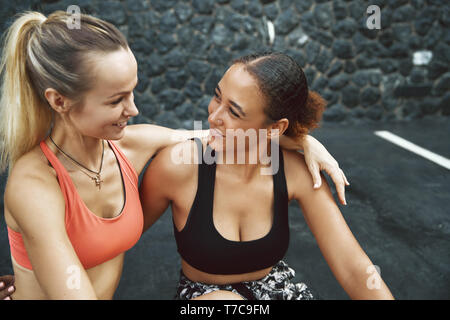 Mettre en place deux jeunes femmes dans les vêtements de sport assis bras dessus bras dessous ensemble dans un parking en dehors de parler et rire après leur course Banque D'Images