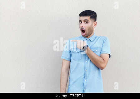 Portrait de choqué beau jeune homme barbu en chemise bleue debout, looking at camera avec surprise le visage et pointant copyspace. Piscine studio Banque D'Images