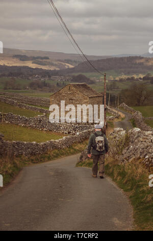 Man de marcher seul dans une petite route de campagne avec un sac à dos et une ancienne grange en pierre, Wharfedale, Yorkshire, UK Banque D'Images