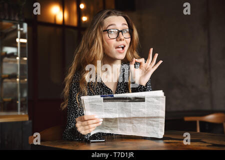 Image d'une belle jeune femme assise dans le café à l'intérieur du journal de lecture montrant bon geste. Banque D'Images