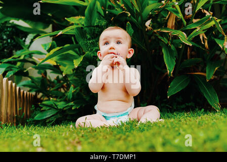 Bébé en couche sans vêtements assis sur l'herbe de grignoter une fleur. Banque D'Images