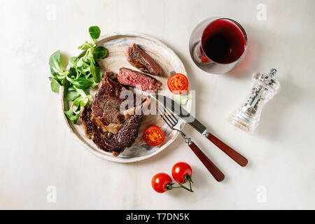 Tranches de rôti grillé à point Pavé de boeuf servi dans une plaque en céramique avec champ vert salade, tomates cerise, couverts, poivre, verre de vin rouge sur Banque D'Images