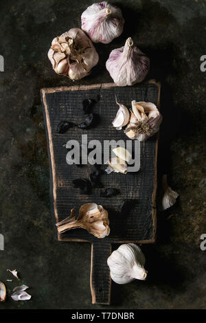 Variété de bulbes bio frais et entiers pelés et gousses d'ail fermenté noir sur planche en bois sombre sur fond de métal. Télévision Banque D'Images