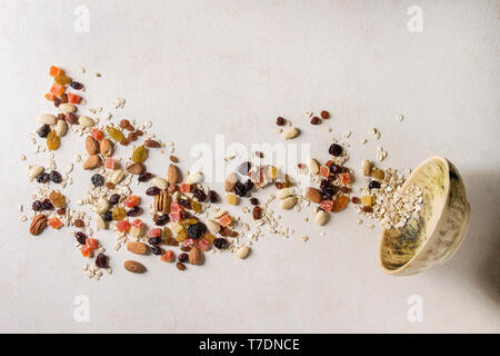 Variété de fruits secs, noix et flocons d'avoine se déverse dans le bol pour la cuisson en céramique petit-déjeuner sain composé de muesli ou granola barres énergétiques sur tex blanc Banque D'Images