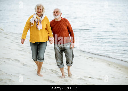 Joli couple vêtus de chandails colorés marche sur la plage de sable, en profitant du temps libre pendant la retraite près de la mer Banque D'Images