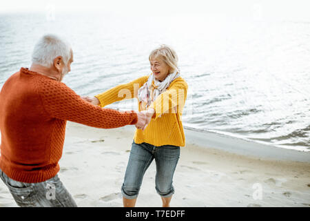 Joli couple vêtus de chandails colorés dansant sur la plage de sable, en profitant du temps libre pendant la retraite près de la mer Banque D'Images