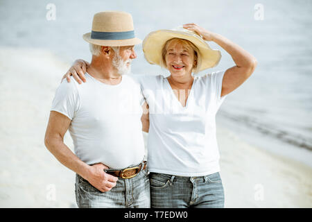 Happy senior couple habillé en blanc T-shirts et des chapeaux à marcher ensemble sur la plage de sable au cours de leur retraite Banque D'Images