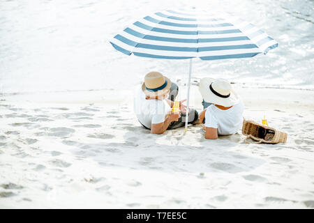 Senior couple sitting ensemble sous parasol sur la plage de sable, profitant de leur retraite près de la mer, vue arrière Banque D'Images