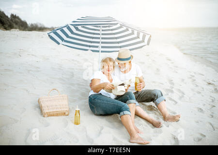 Happy senior couple relaxing, couché ensemble sous parasol sur la plage de sable, profitant de leur retraite près de la mer Banque D'Images