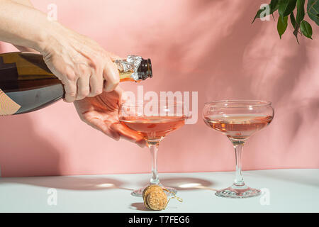 La main féminine pouring champagne ou vin dans des verres. Fond rose doux plein soleil. Espace copie le minimalisme. Banque D'Images