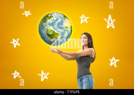 Young smiling brunette girl wearing jean décontracté et t-shirt avec les paumes vers le haut, earth globe et personnages tirés sur fond jaune Banque D'Images