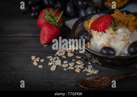 Mélanger le yogourt gruau, fraise et raisin topping en noir sur bois bol avec cuillère de table, raisin, fraise et le gruau placés autour. Banque D'Images