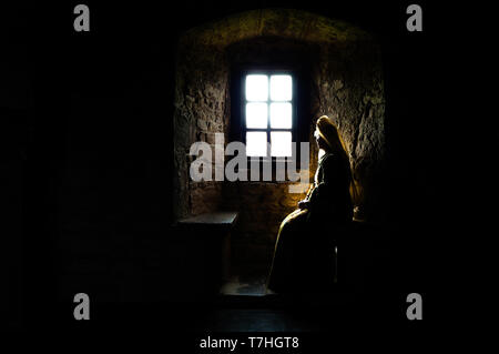 Silhouette partielle d'une femme en robe médiévale assis dans le noir à la recherche d'une fenêtre lumineuse dans les profondeurs d'un ancien château Banque D'Images