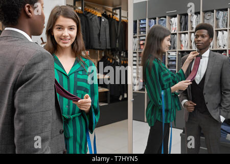 Jeune homme africain debout dans boutique, à la recherche de miroir, le choix d'une cravate. Girl shop médecin client de choisir une cravate et souriant. Homme portant un costume gris et en chemise blanche. Banque D'Images