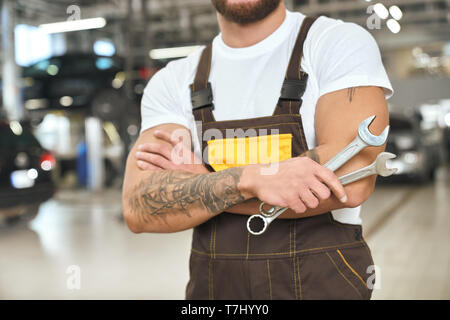 Mécanicien musculaire méconnaissable avec mains tattoed, vêtu d'une combinaison et en t shirt blanc. L'homme brutal holding clés, travaillant en autoservice. Banque D'Images