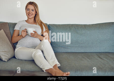 Jolie jeune femme enceinte pieds nus sur un canapé relaxant de boire une tasse de thé chaud et souriant joyeusement à l'appareil photo Banque D'Images